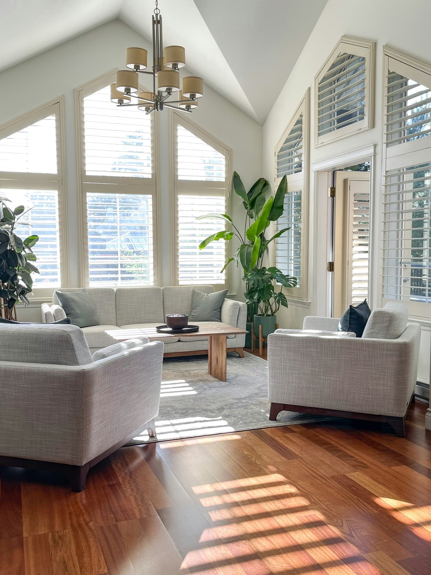 A sunlit living room with vaulted ceilings, plantation shutters, light sofas and hardwood floors